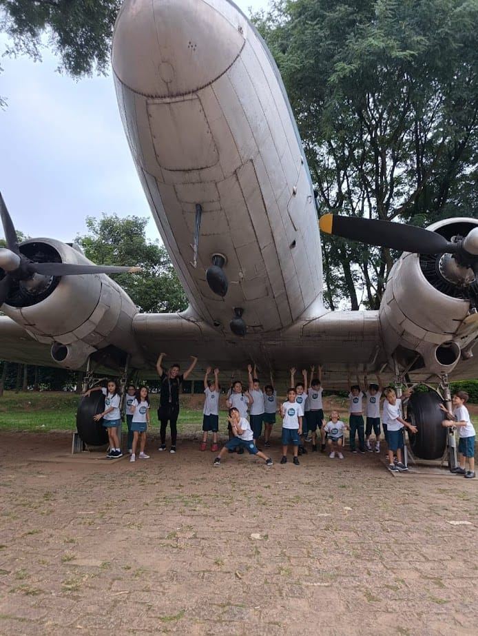Grupo de estudantes uniformizados posando sob um avião antigo em passeio pedagógico