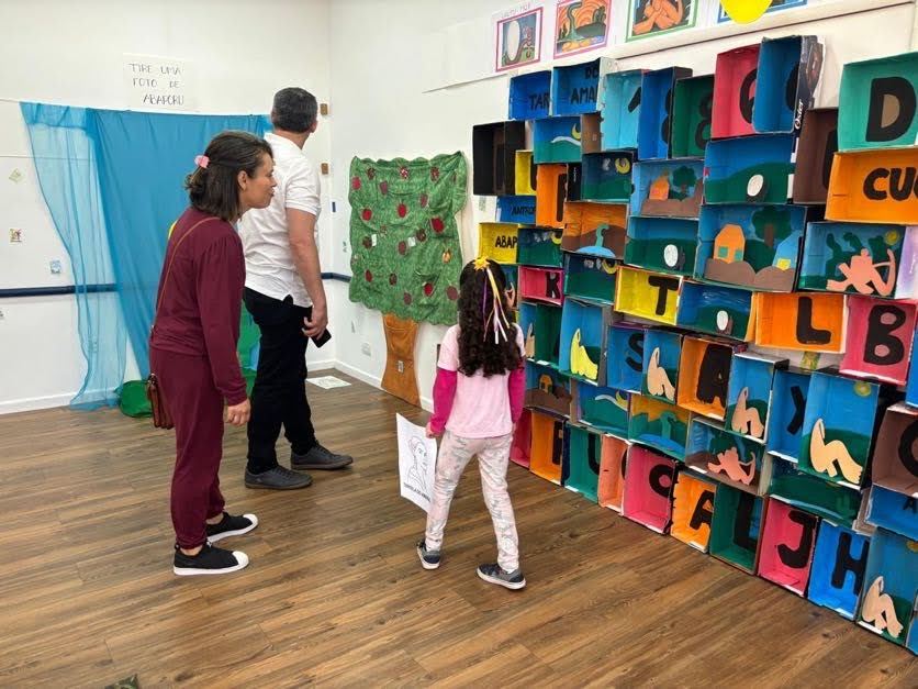 Criança observando uma parede colorida de trabalhos artísticos com letras, durante mostra de artes na escola