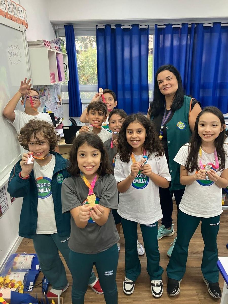 Estudantes sorridentes exibindo medalhas de olimpíada ao lado de uma professora