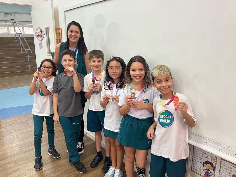 Estudantes uniformizados e uma professora em sala de aula, mostrando medalhas de olimpíada