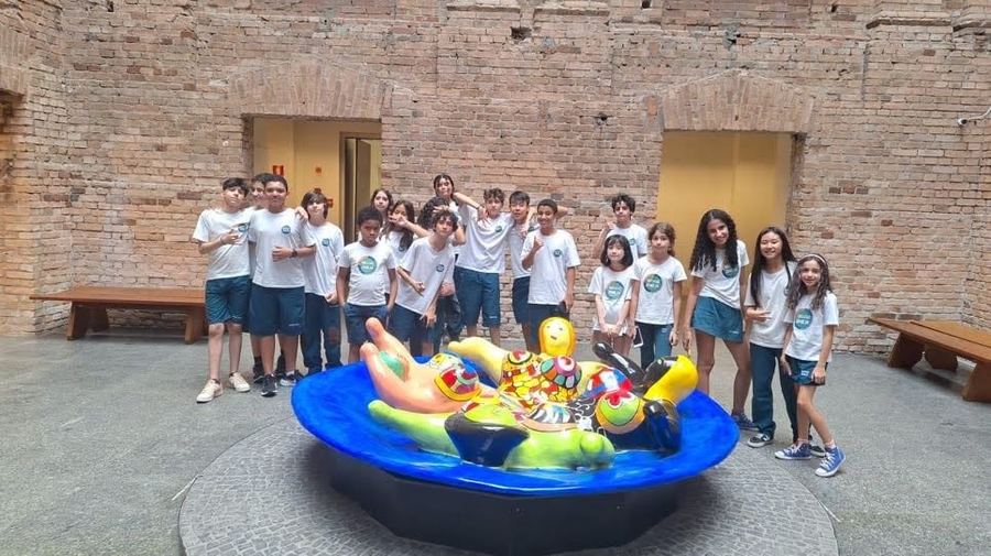Estudantes uniformizados em pátio de museu, ao redor de uma escultura colorida